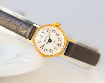 Peut inclure: Une montre-bracelet dorée avec un cadran blanc et un bracelet en cuir noir. Le cadran de la montre comporte des chiffres romains noirs et le mot "Чайка" (Chaika) imprimé en noir.
