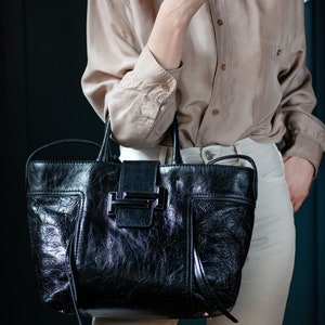 A black leather Tod's tote bag with a silver buckle closure. The bag has a textured leather finish and a long strap. Features a prominent tonal Double T logo leather appliqué clasp closure across the front tab.I ncludes a secondary long, flat leather shoulder strap for versatile styling.