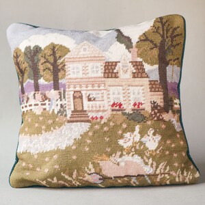 May include: A green and white needlepoint pillow featuring a country house with a white picket fence, trees, and birds. The house has a brown roof and white trim.