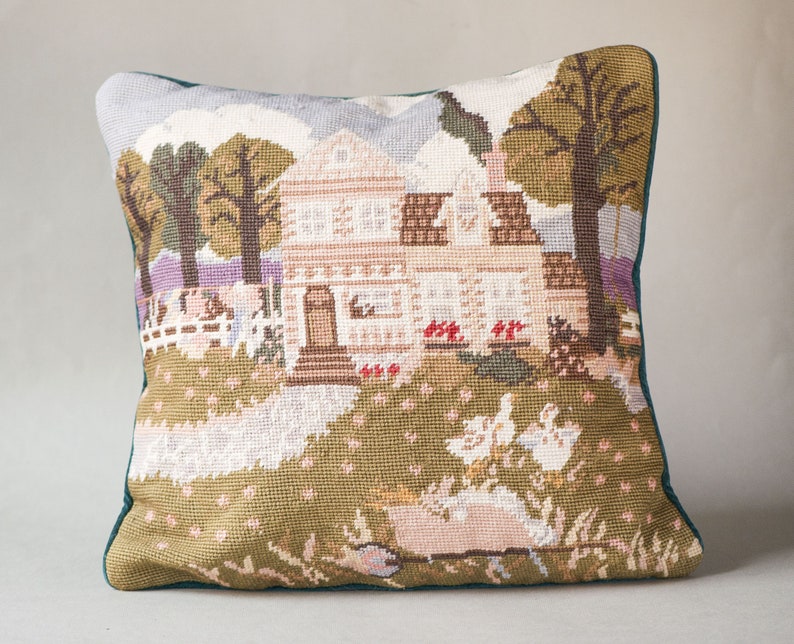 May include: A green and white needlepoint pillow featuring a country house with a white picket fence, trees, and birds. The house has a brown roof and white trim.