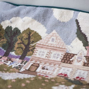 May include: A green and brown needlepoint pillow with a scene of a house with a white roof, trees, and a fence. The house has brown trim and windows. The background is a light blue sky with white clouds.