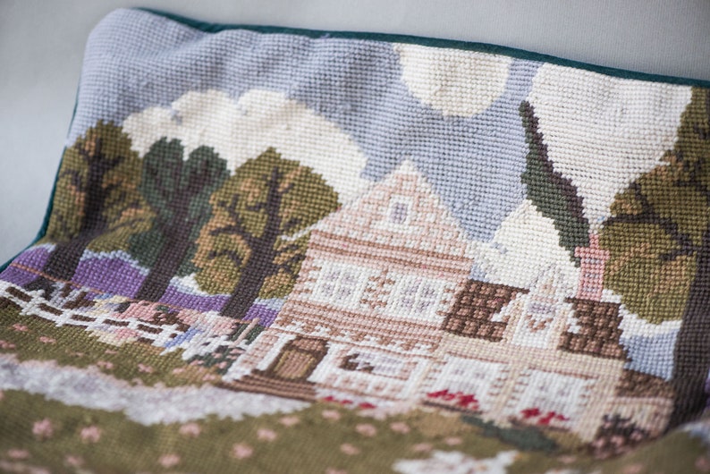 May include: A green and brown needlepoint pillow with a scene of a house with a white roof, trees, and a fence. The house has brown trim and windows. The background is a light blue sky with white clouds.