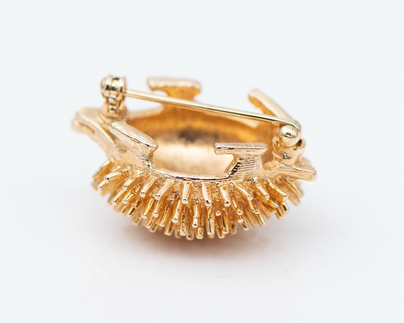 May include: A gold-toned brooch in the shape of a hedgehog. The hedgehog is made of metal and has a textured surface. The brooch has a pin on the back for attaching it to clothing.