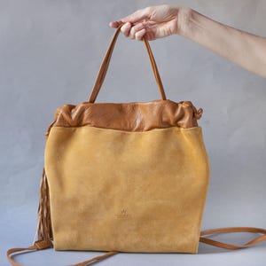 Puede incluir: Bolso de mano de dos tonos con un cuerpo de ante amarillo mostaza y una parte superior y asas de cuero marrón. El bolso tiene un cierre de cordón y una correa larga para el hombro. Una borla cuelga del lateral. El bolso está sostenido por una mano.