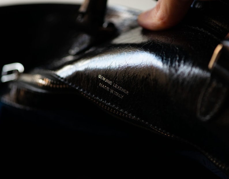 Op de afbeelding: Close-up van een zwarte leren tas met een zilveren rits en de tekst "Genuine Leather Made in Italy" gegraveerd aan de binnenkant.