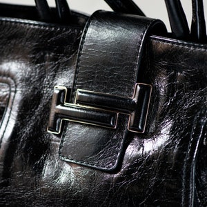 Op de afbeelding: Close-up van een zwarte leren handtas met een goudkleurige T-vormige sluiting.