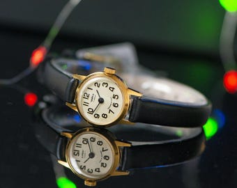 Peut inclure: Une montre-bracelet dorée avec un cadran rond crème, des chiffres arabes noirs et de fines aiguilles noires. La montre possède un bracelet en cuir noir et une petite couronne sur le côté. Le cadran porte l'inscription Chaika en cyrillique.
