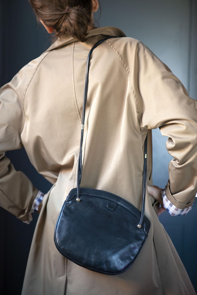 Puede incluir: Un bolso bandolera de cuero negro con forma curvada, colgado de una fina correa negra. El bolso se lleva sobre una gabardina beige. El abrigo tiene un dise&ntilde;o cl&aacute;sico con cuello y cierre de botones. Las mangas est&aacute;n dobladas, revelando un estampado de cuadros.