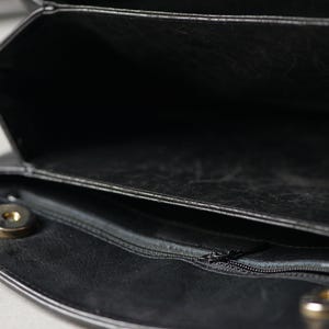 Puede incluir: Primer plano de un bolso de mano de cuero negro con m&uacute;ltiples compartimentos. El bolso tiene un cierre de solapa con un broche magn&eacute;tico dorado. Una cremallera es visible en el interior, y el bolso parece estar abierto.