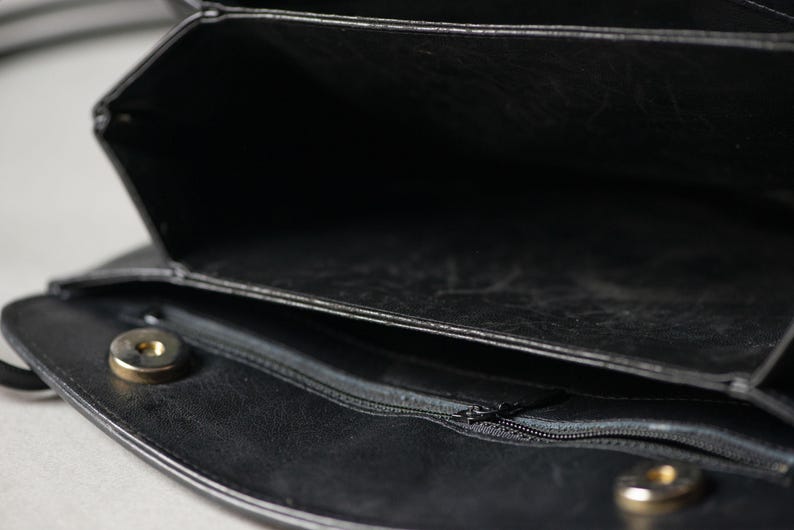 Puede incluir: Primer plano de un bolso de mano de cuero negro con m&uacute;ltiples compartimentos. El bolso tiene un cierre de solapa con un broche magn&eacute;tico dorado. Una cremallera es visible en el interior, y el bolso parece estar abierto.