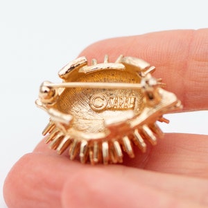 May include: Gold-toned brooch with a sunburst design. The back of the brooch has a circular, gold-toned backing with the word "SARAH" engraved in the center.