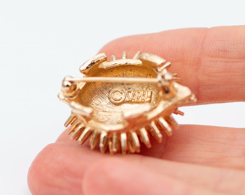 May include: Gold-toned brooch with a sunburst design. The back of the brooch has a circular, gold-toned backing with the word "SARAH" engraved in the center.