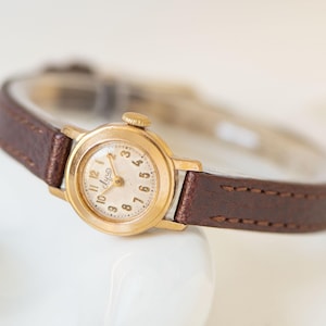 Könnte beinhalten: Eine goldfarbene Vintage-Armbanduhr mit einem braunen Lederarmband. Das Zifferblatt der Uhr ist cremefarben mit schwarzen Ziffern und Zeigern. Das Armband ist etwa 1,3 cm breit. Ein klassisches Accessoire.