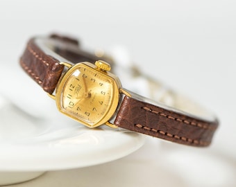Peut inclure: Une montre-bracelet analogique dorée avec un boîtier carré et un bracelet en cuir marron texturé. Le cadran présente des chiffres noirs, des aiguilles dorées et le texte Zaria 17 jewels Made in USSR imprimé en noir sur le cadran.