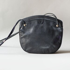 Puede incluir: Un bolso bandolera de cuero negro con forma redondeada. El bolso tiene una correa larga y fina y peque&ntilde;os detalles dorados. El bolso est&aacute; hecho de cuero liso y mate.
