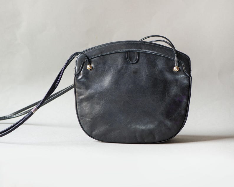 Puede incluir: Un bolso bandolera de cuero negro con forma redondeada. El bolso tiene una correa larga y fina y peque&ntilde;os detalles dorados. El bolso est&aacute; hecho de cuero liso y mate.