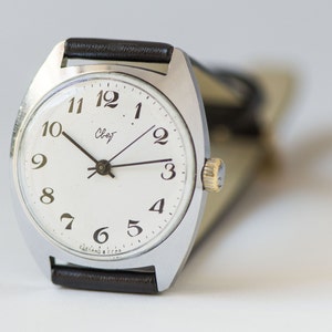 May include: A silver wristwatch with a white face and black hands. The watch has black Roman numeral hour markers and a black leather band.