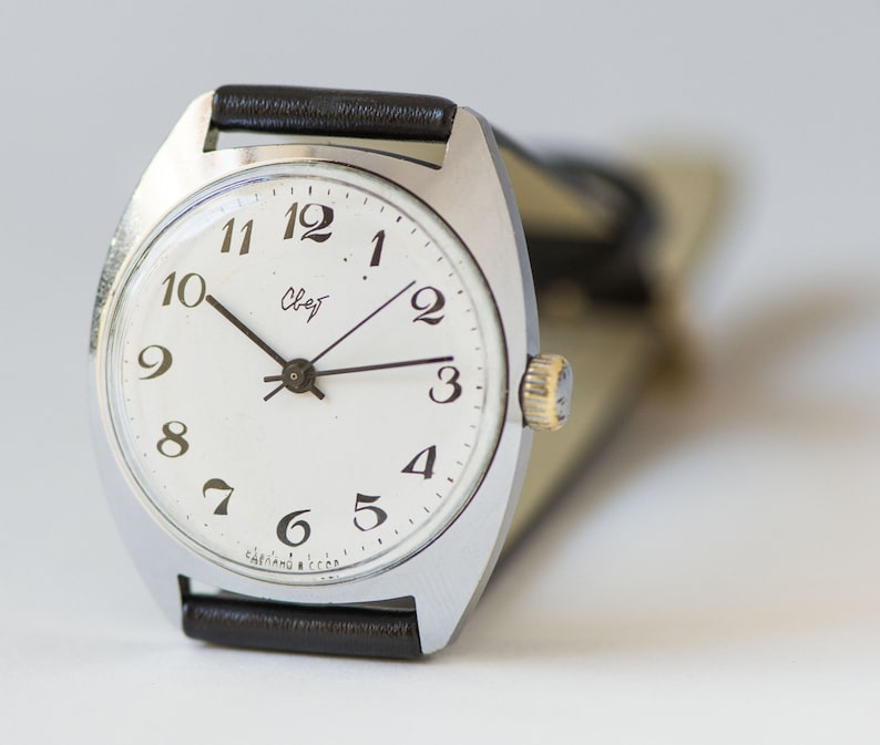 May include: A silver wristwatch with a white face and black hands. The watch has black Roman numeral hour markers and a black leather band.