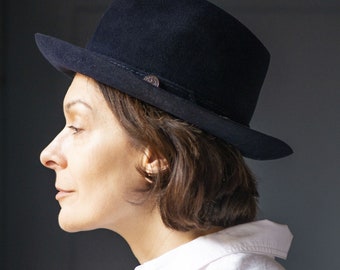 May include: A woman wearing a black fedora hat with a white button-down shirt.