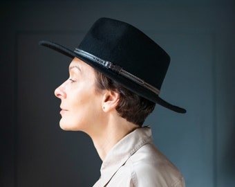 May include: A person wearing a black fedora hat with a leather band. The hat has a wide brim and is angled slightly to the side.