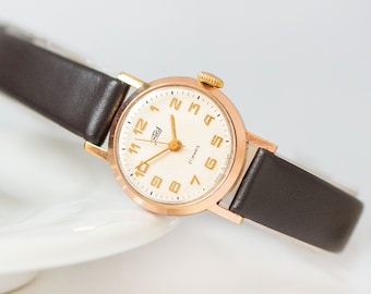 Peut inclure: Une montre-bracelet dorée avec un cadran blanc et un bracelet en cuir noir. Le cadran de la montre comporte des chiffres romains noirs et une aiguille des secondes dorée. Le cadran de la montre comporte également le texte "21 jewels" et "Made in USSR".