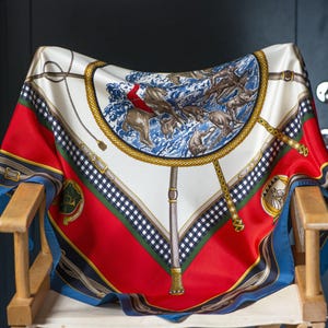 Può includere: Un foulard di seta bianco con un motivo geometrico rosso, blu e nero. Il centro del foulard presenta un design circolare con una catena dorata e un motivo a onde blu e bianco. Il foulard è drappeggiato su una sedia di legno.