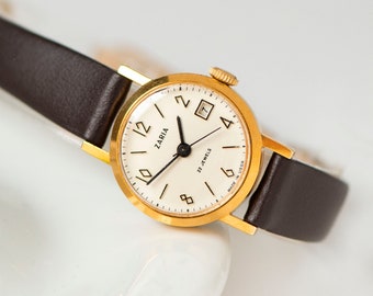 Peut inclure: Une montre-bracelet Zaria dorée avec un cadran blanc, des aiguilles noires et des chiffres romains noirs. La montre a un bracelet en cuir marron et indique "22 Jewels" et "Made in USSR" sur le cadran.