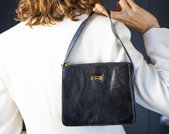May include: A black leather handbag with a rectangular silhouette, a single shoulder strap, and a gold-toned metal rectangular accent piece centered on the front. The bag features a top zipper closure with a gold-toned metal pull tab.