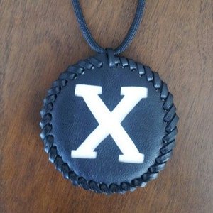 May include: A black leather pendant necklace featuring a large white letter "X". The pendant is round with a braided edge and hangs from a black cord. The background is a dark wood surface.