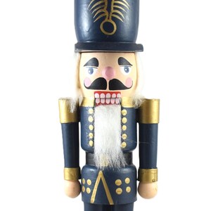 Puede incluir: Figura de cascanueces de madera con uniforme azul marino y detalles dorados. La figura tiene barba blanca, boca roja y bigote negro. El sombrero tiene un emblema dorado. El cascanueces está sobre una base azul marino.