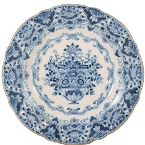 Può includere: Un piatto rotondo in ceramica con bordo smerlato, caratterizzato da una base bianca e complessi motivi floreali blu. Il centro mostra un'urna dettagliata piena di fiori, circondata da un bordo decorativo di fiori e fogliame stilizzati.