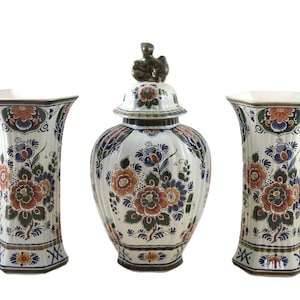 Op de afbeelding: Een set van drie keramische vazen met een bloemenontwerp. De set bevat twee hoge vazen en een pot met deksel. De vazen en de pot zijn wit met een herhalend patroon van oranje, blauwe en groene bloemen.