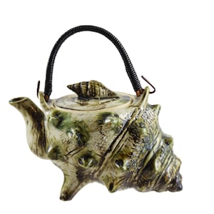 May include: A ceramic teapot shaped like a conch shell, with a green and cream glaze. The teapot has a black woven handle and a shell-shaped lid finial. The spout and base are also shell-like in design. A unique piece for tea lovers.