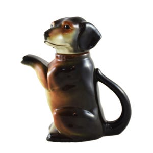 Puede incluir: Una tetera de cerámica con forma de perro salchicha en posición sentada, con la pata delantera levantada. La tetera es principalmente marrón y negra, con detalles en beige y crema en la cabeza y el pecho. El asa es un lazo negro curvado.