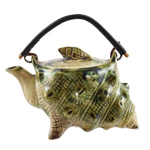 May include: A ceramic teapot shaped like a conch shell, with a green and beige glaze. The teapot has a black woven handle and a small shell-shaped lid. The design features textured details and a spout for pouring.