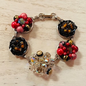 Puede incluir: Un brazalete vintage con una cadena plateada y eslabones circulares decorativos. Cada eslabón presenta grupos de cuentas rojas, rosas, negras y doradas, con detalles de cristal iridiscente. El brazalete tiene un cierre de palanca.