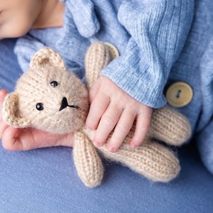 Może przedstawiać: Zbliżenie na rękę dziecka trzymającego brązowego pluszowego misia z dzianiny. Dziecko ma na sobie jasnoniebieski dzianinowy śpioszek.