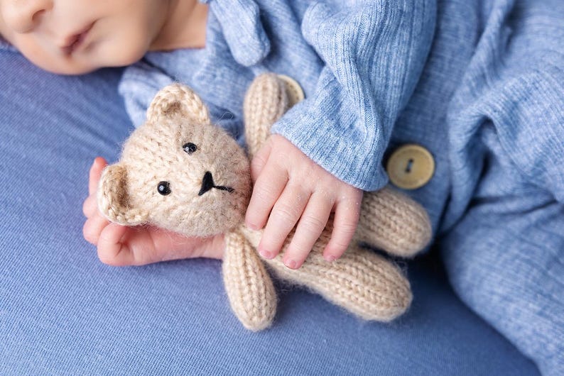 Może przedstawiać: Zbliżenie na rękę dziecka trzymającego brązowego pluszowego misia z dzianiny. Dziecko ma na sobie jasnoniebieski dzianinowy śpioszek.