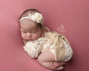 May include: A newborn baby wearing a white lace romper with a large beige bow on the back. The baby is wearing a white headband with a small flower embellishment. The baby is sleeping on a pink background.