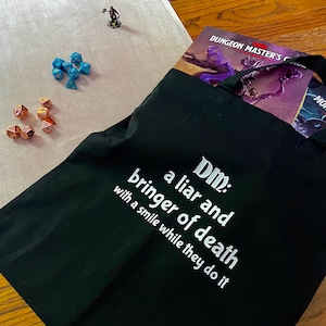 May include: Black tote bag with white text that reads "DM: a liar and bringer of death with a smile while they do it". The bag is filled with Dungeons and Dragons books and dice.