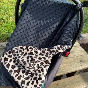May include: A black and white leopard print car seat cover with a black minky dot fabric on the back side. The cover is designed to fit over a car seat and has straps to secure it in place.