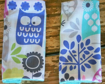 May include: Two fabric dish towels with a white background and colorful patterns. One towel features a blue owl with white eyes and a green leaf design. The other towel features a blue tree with gray and turquoise leaves.