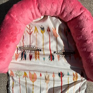 May include: Pink and white baby head support pillow with a pattern of colorful arrows. The pillow is made of soft, plush fabric and has a comfortable shape to support a baby's head and neck.