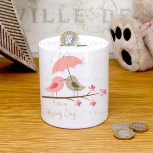 May include: White ceramic money box with a coin slot. The design shows two cartoon birds under a pink umbrella on a branch with pink flowers. The text "For a Rainy Day" is below the birds. A coin is in the slot.