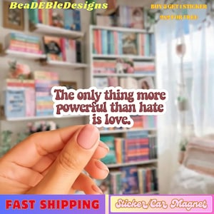 May include: A white sticker with maroon text that reads, "The only thing more powerful than hate is love." The sticker is held in a hand with a blurred background of bookshelves. The sticker is a car magnet.