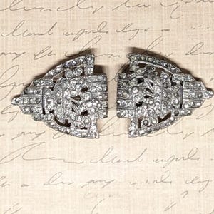 May include: Two silver-toned, Art Deco style clasps, each encrusted with small, clear rhinestones. The clasps are open, revealing their intricate floral and geometric designs. The background is a cream-colored paper with handwritten script.