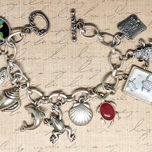 May include: A silver charm bracelet with a variety of sea-themed charms, including a starfish, a seashell, a seahorse, a dolphin, a frog, a turtle, and a red stone. The bracelet also features a charm with the text "TRUST YOUR GUT FEELING".