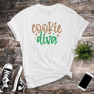 May include: White t-shirt with the text "cookie diva" in brown and green lettering with small gold stars.