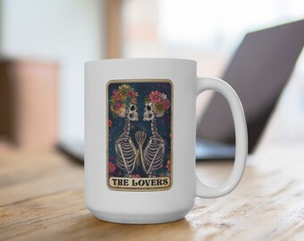 May include: White ceramic coffee mug with a colorful illustration of two skeletons wearing floral crowns. The skeletons are holding hands and the text "THE LOVERS" is printed below them.