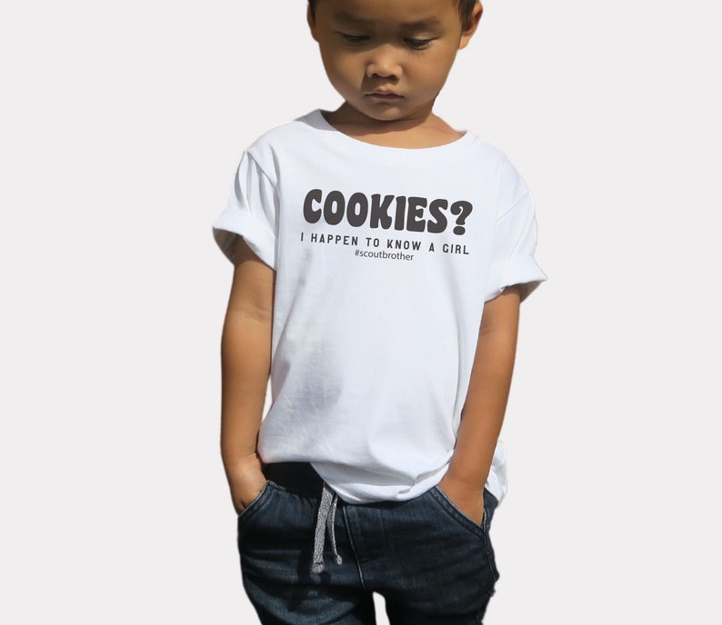May include: A white t-shirt with the text "COOKIES? I HAPPEN TO KNOW A GIRL #scoutbrother" in black. The shirt is worn by a young child, paired with blue jeans.